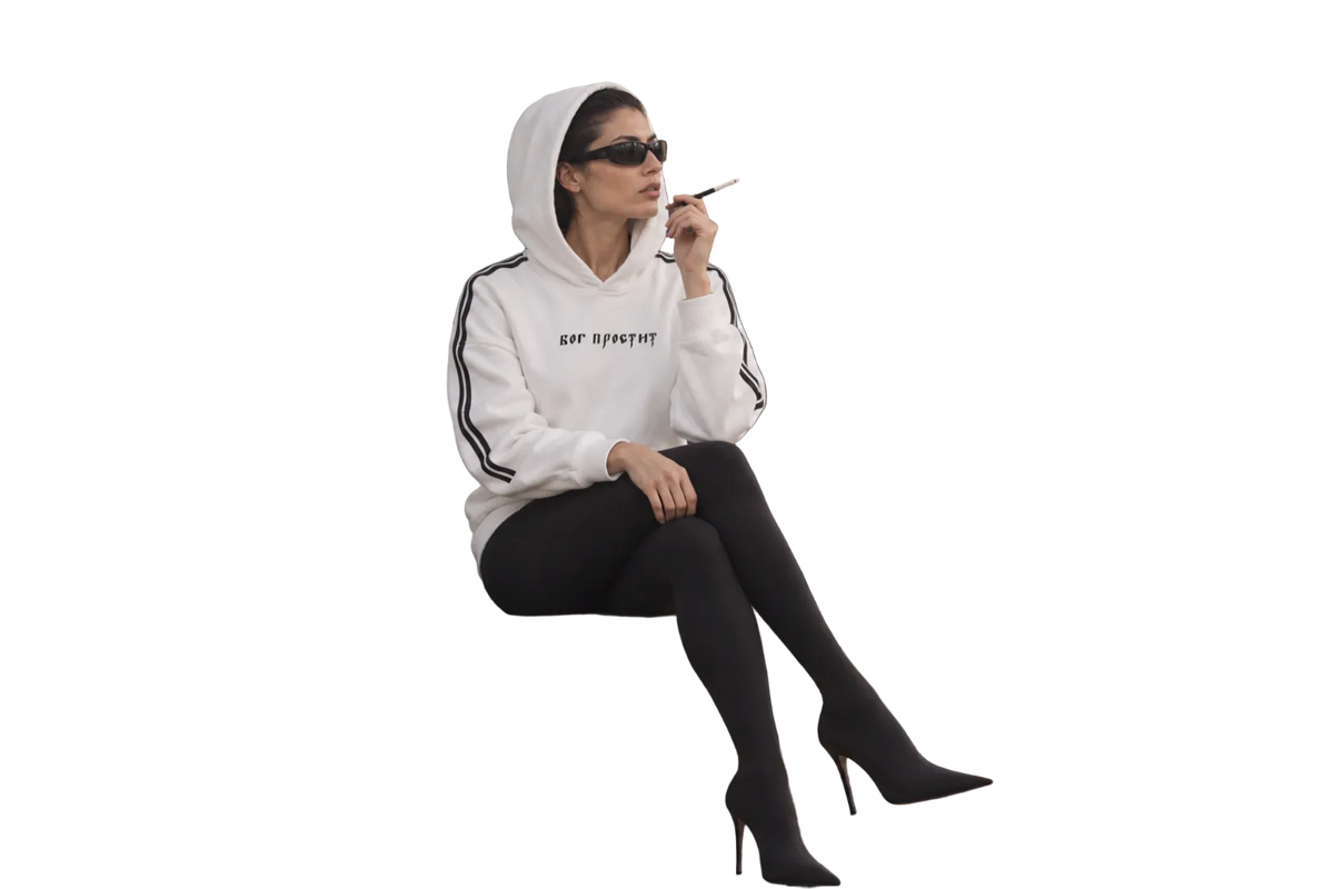 Seated model wearing a white God Forgives hoodie with black sleeve stripes, black tights, sunglasses and heeled boots